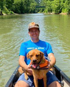 在森林环绕的河上，穿着“徒步游”t恤的男子和穿着救生衣的狗在皮划艇上微笑.